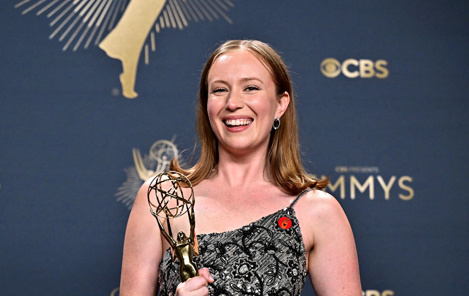 Hannah Einbinder holding her Emmy with a floral silver dress and red Artists4Ceasefire pin.