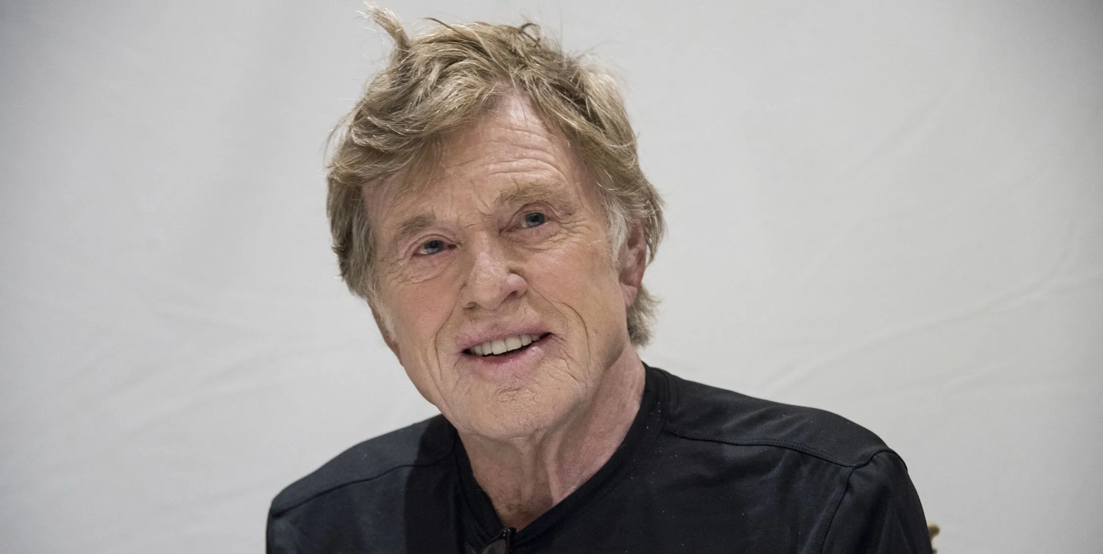 Robert Redford smiles while wearing a black shirt and sitting against a grey background.