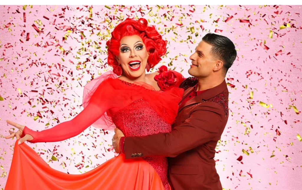 La Voix with red hair and red sequined earings and dress being held by her dance partner, Aljaz Škorjanec, in a maroon suit.