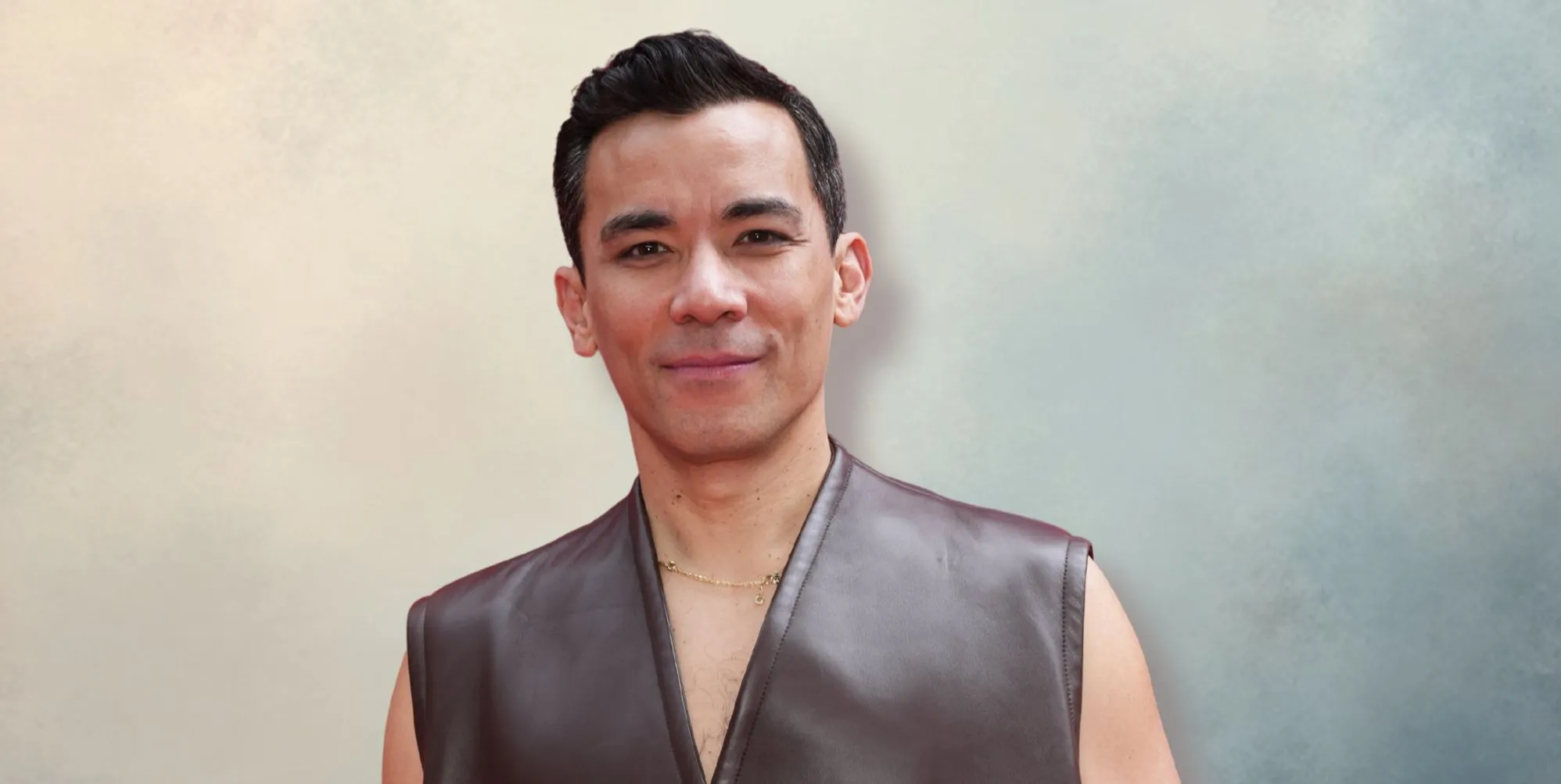 Conrad Ricamora in a grey sleeveless jacket smiling and posing. He is against a blue/grey/cream background