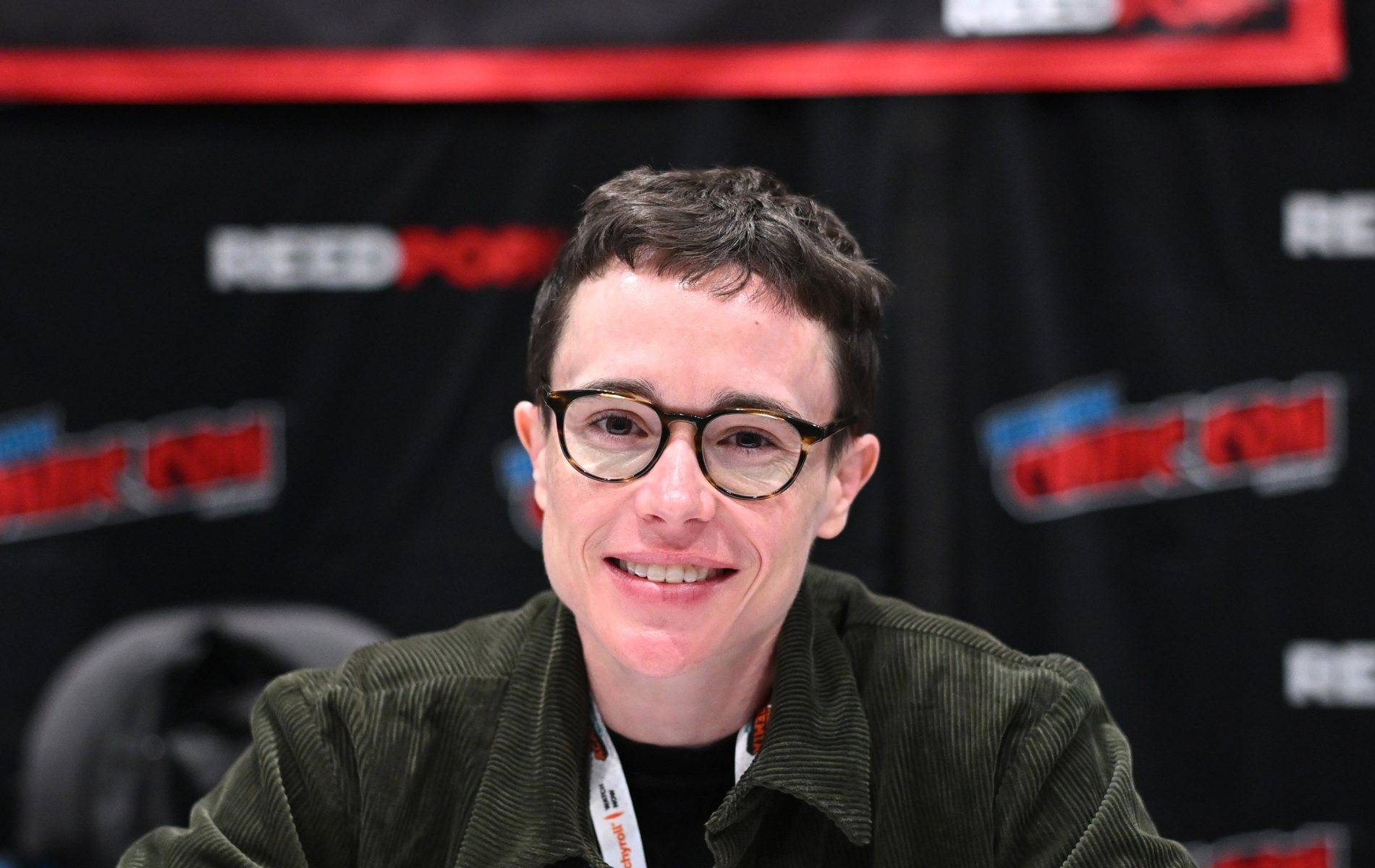 Elliot Page attends New York Comic Con. Eliott wearing a green cordoroy jacket smiling into the camera, wearing round glasses.