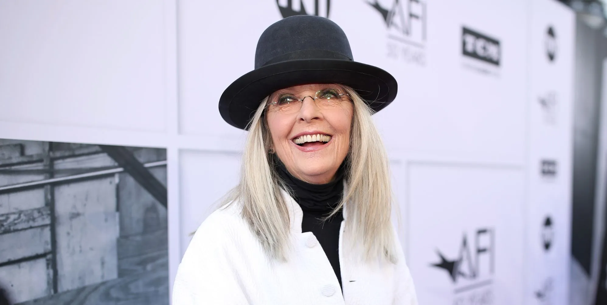 Diane Keaton in a white jacket and black hat laughing.