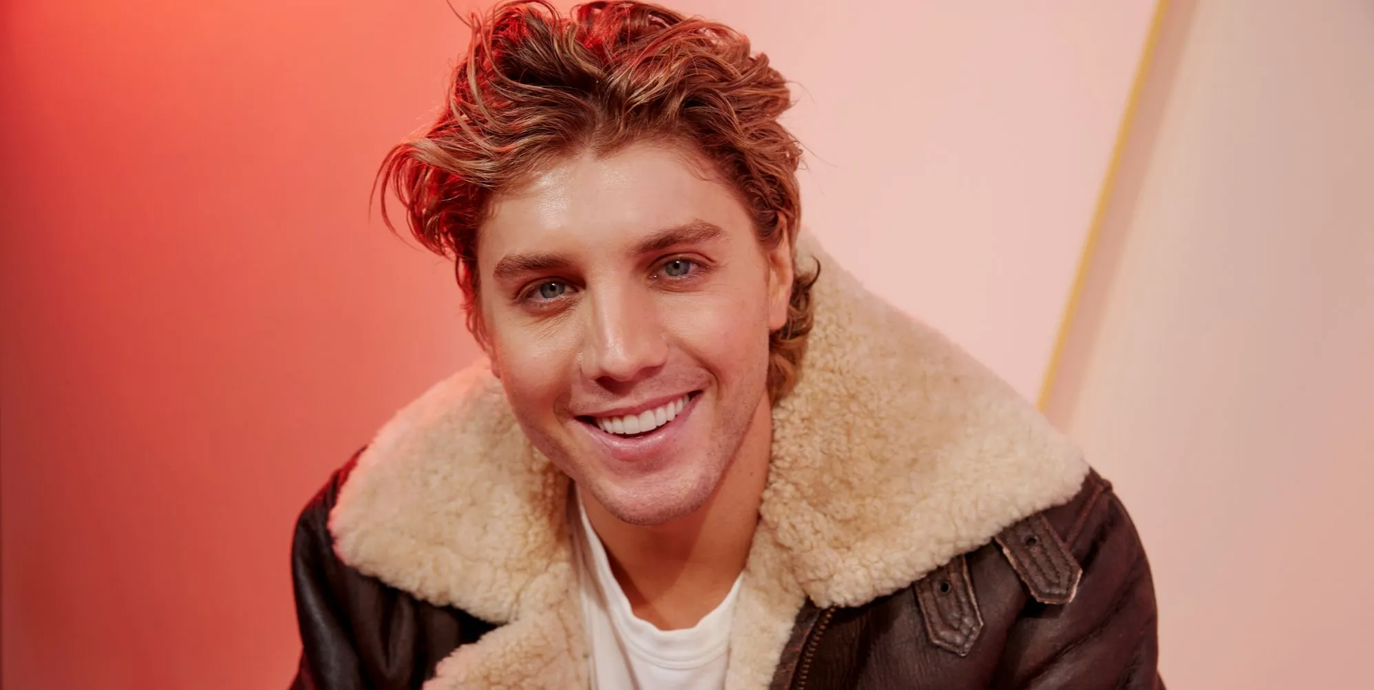 Lukas Gage in a brown leather jacket smiling, sat against an orange background
