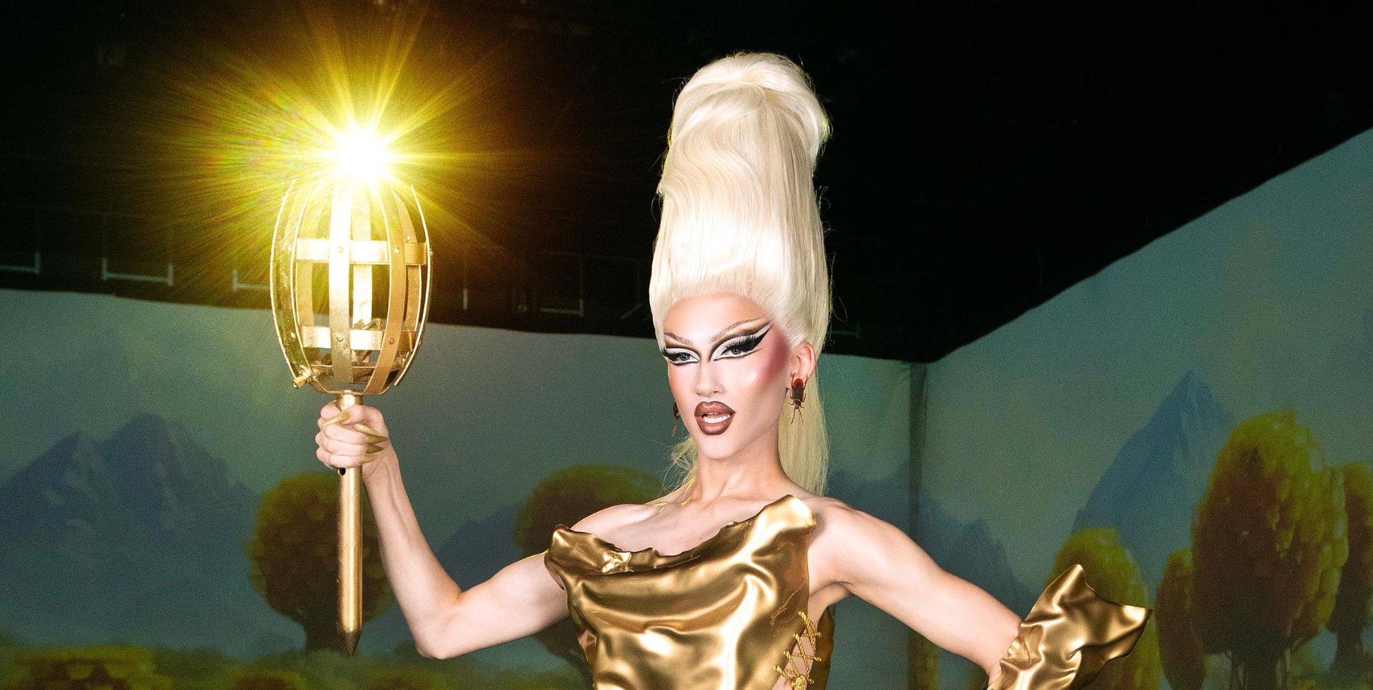 Kyran Thrax in a gold outfit and huge blonde wig with her mouth open. She is holding a gold torch and standing in a grey room.