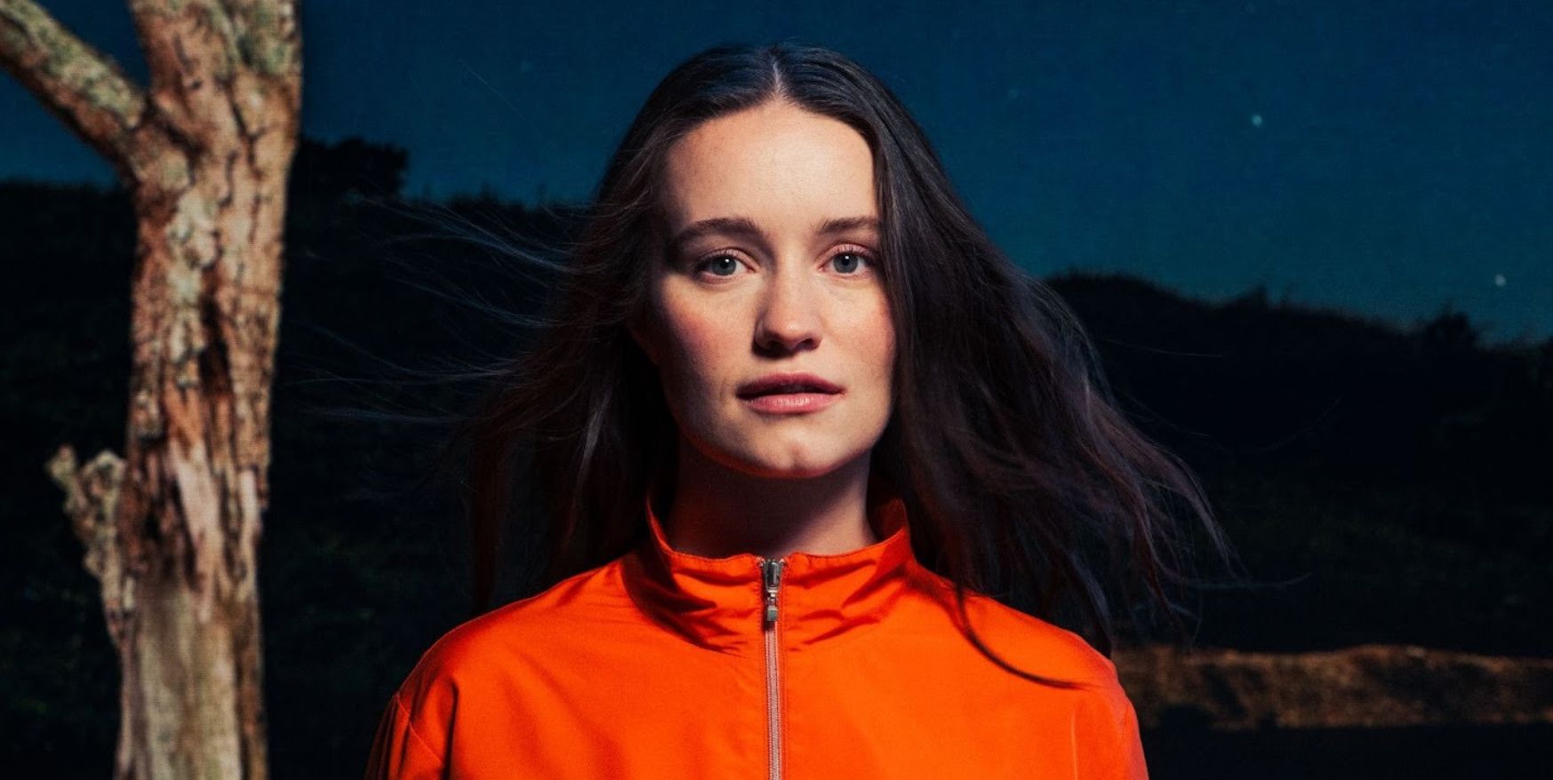Sigrid looking at the camera, wearing an orange zip neck jumper.