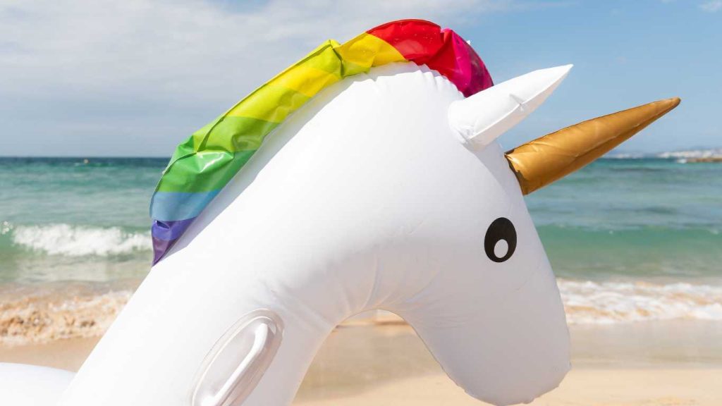 White inflatable unicorn float with rainbow mane and gold horn rests on a sandy beach with ocean waves in the background