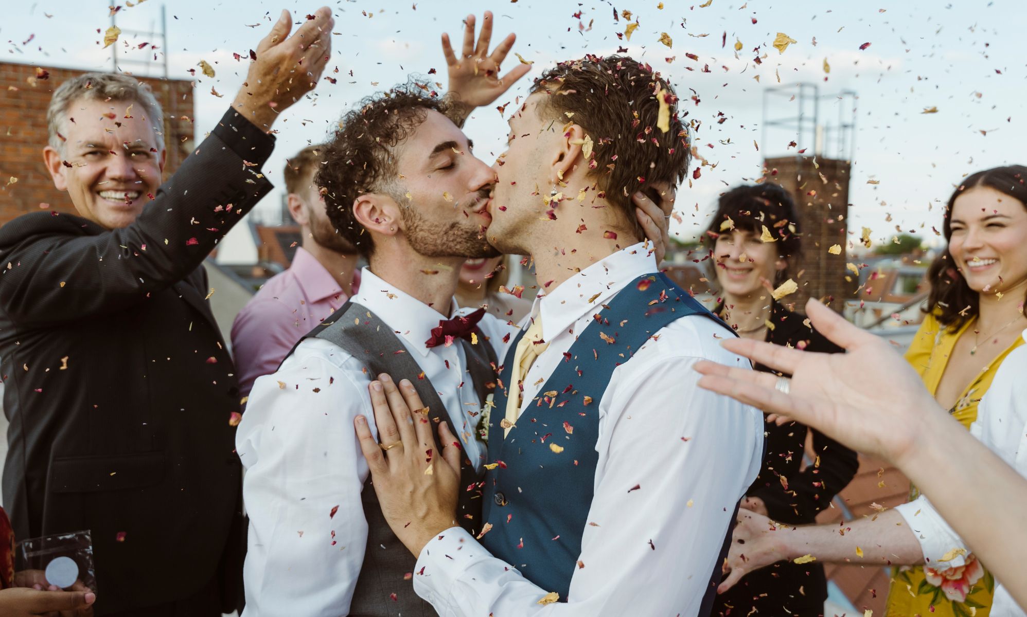 Two men kiss surrounded by a crowd of people throwing confetti.