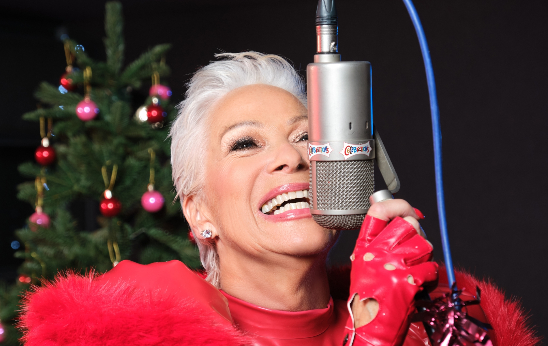 Denise Welch in all red, smiling close to a microphone. Christmas tree in the background