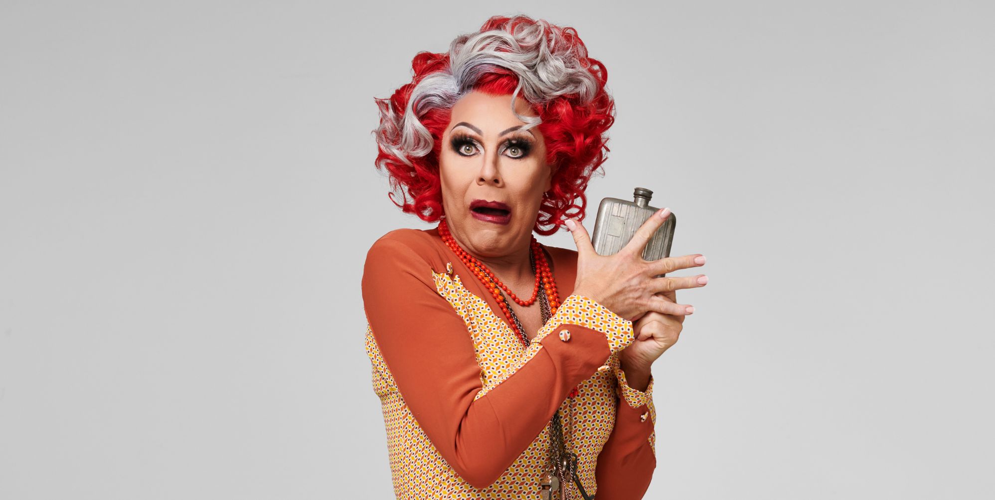 La Voix in an orange top and dress, red and silver wig, holding a whisky bottle and pulling a face.