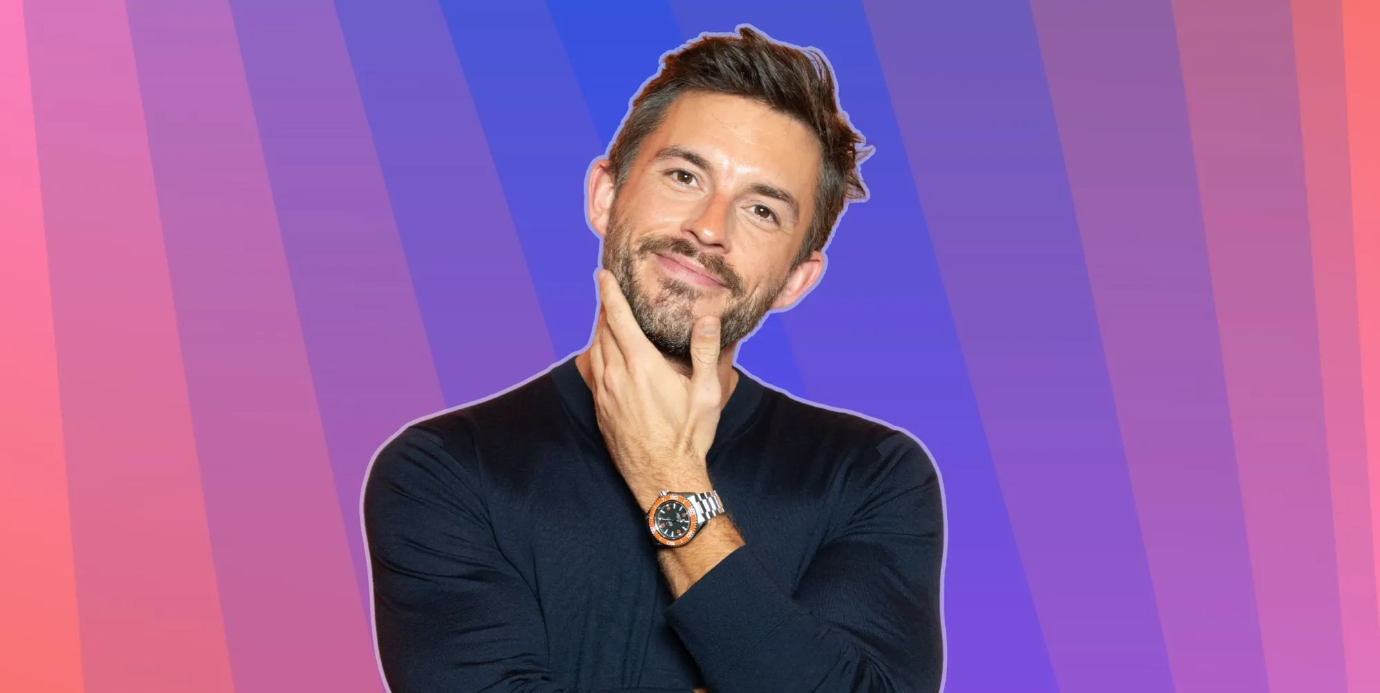 Jonathan Bailey smiles and places his hand on his chin. He is against a multi-coloured background.
