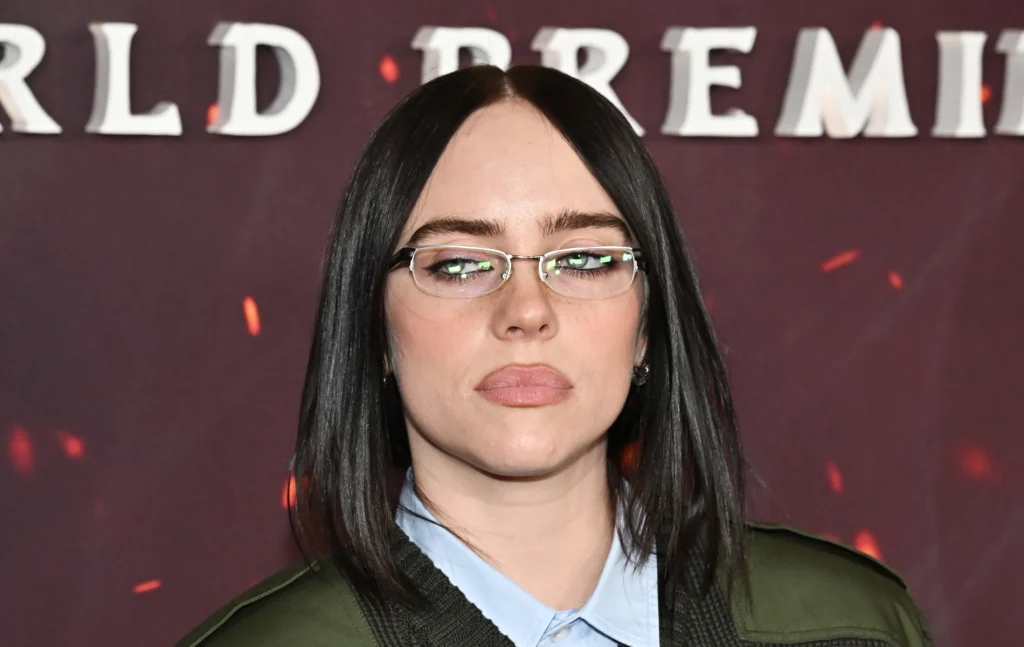 Billie Eilish in small glasses, a light blue shirt and green jacket