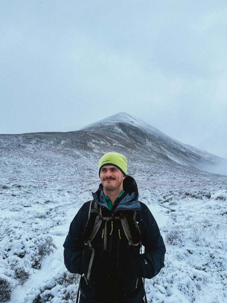 Matthew con un gorro de neón y equipo de senderismo contra una colina nevada como telón de fondo