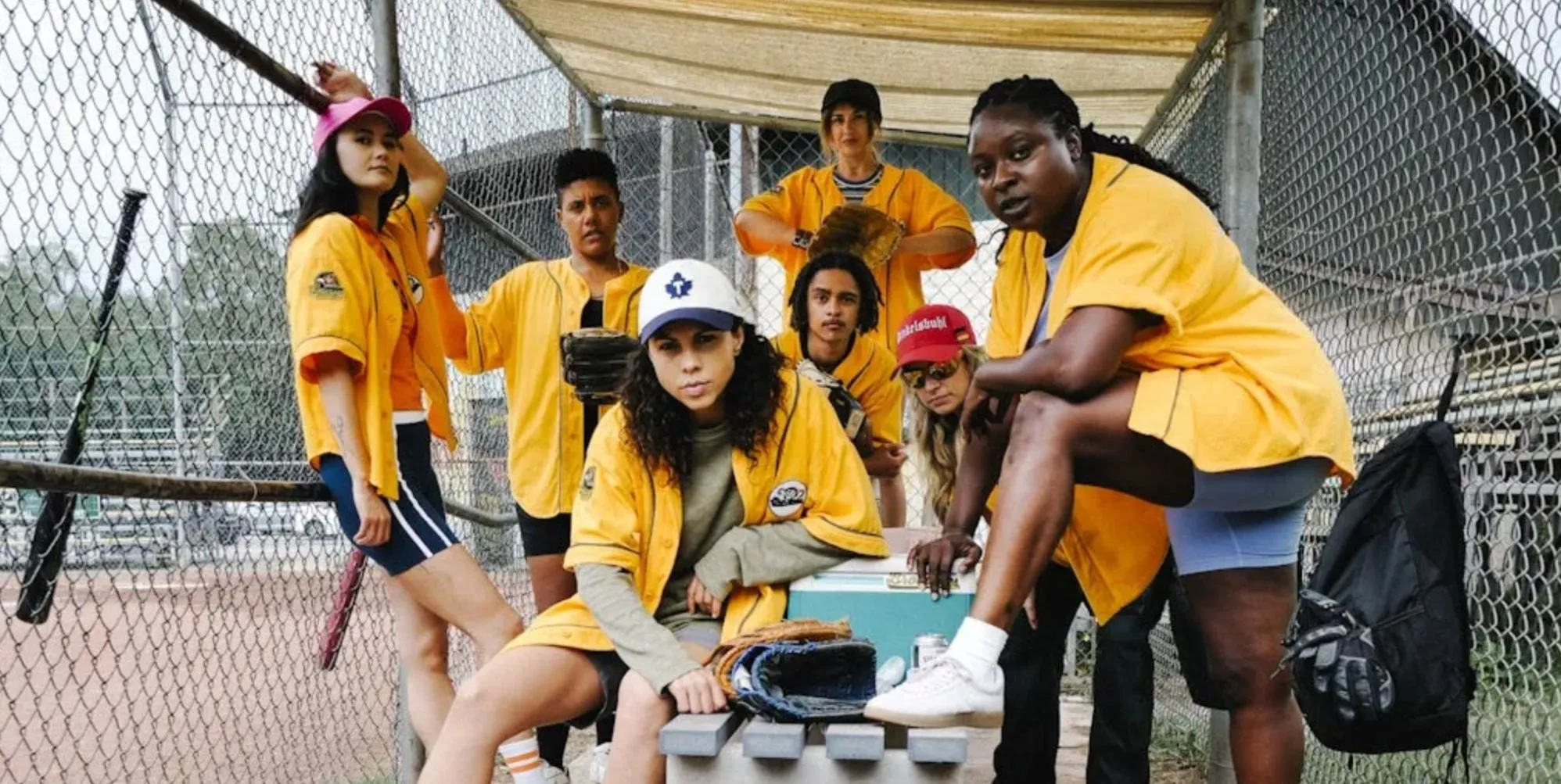 A first look still from upcoming show Slo Pitch showing softball players posing in yellow tops, posing by a pitch, looking at the camera.