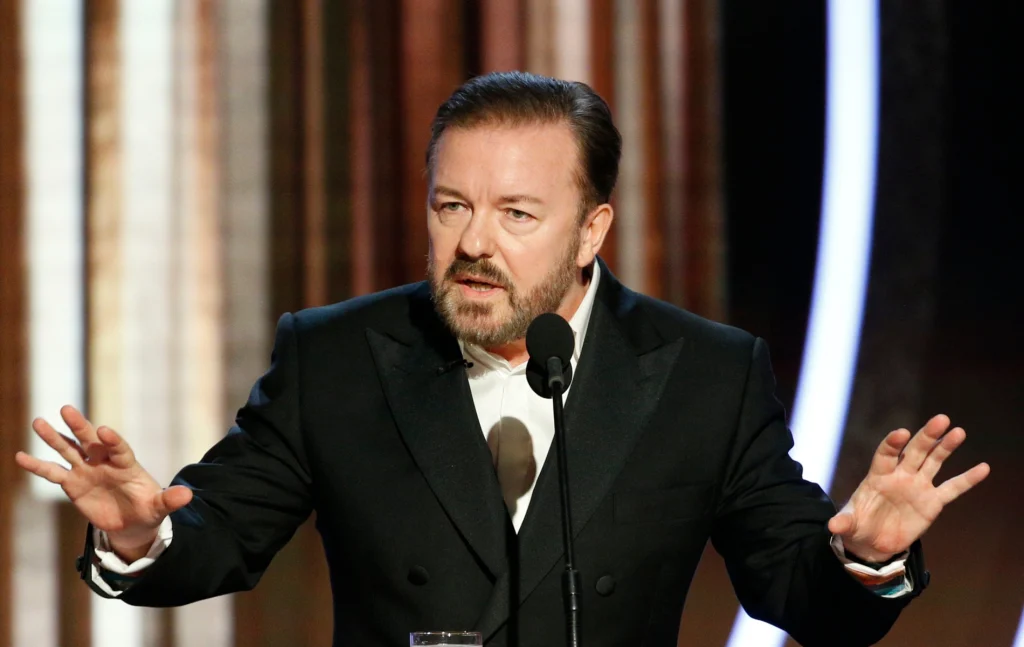 Ricky Gervais speaking into a mic with his hands up
