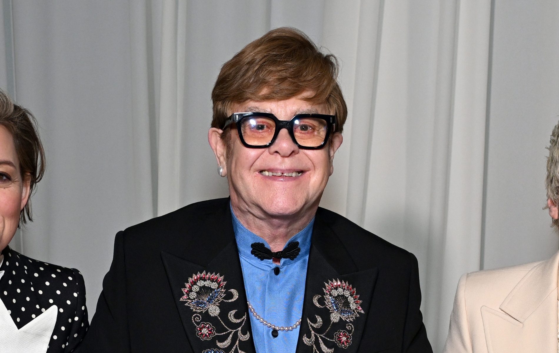 Elton John wearing a blue shirt and black suit jacket with floral embroidery