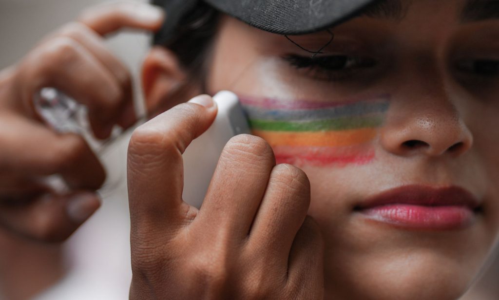 A close-up of someone putting rainbow face paint on their cheek.