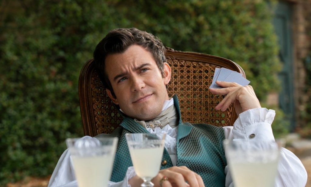 Benedict Bridgerton sitting in a wooden chair with cards and drinks in front of him