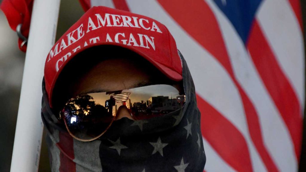 A man wearing a MAGA hat and carrying a flag protests during a pro Trump rally.