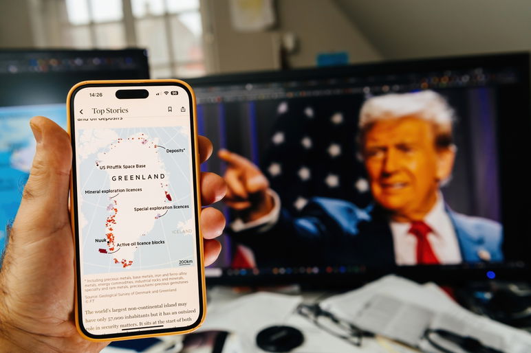 Una mano sosteniendo un teléfono inteligente que muestra un mapa de recursos de Groenlandia con detalles de exploración, mientras dos monitores muestran a Donald Trump señalando y un mapa de Groenlandia.