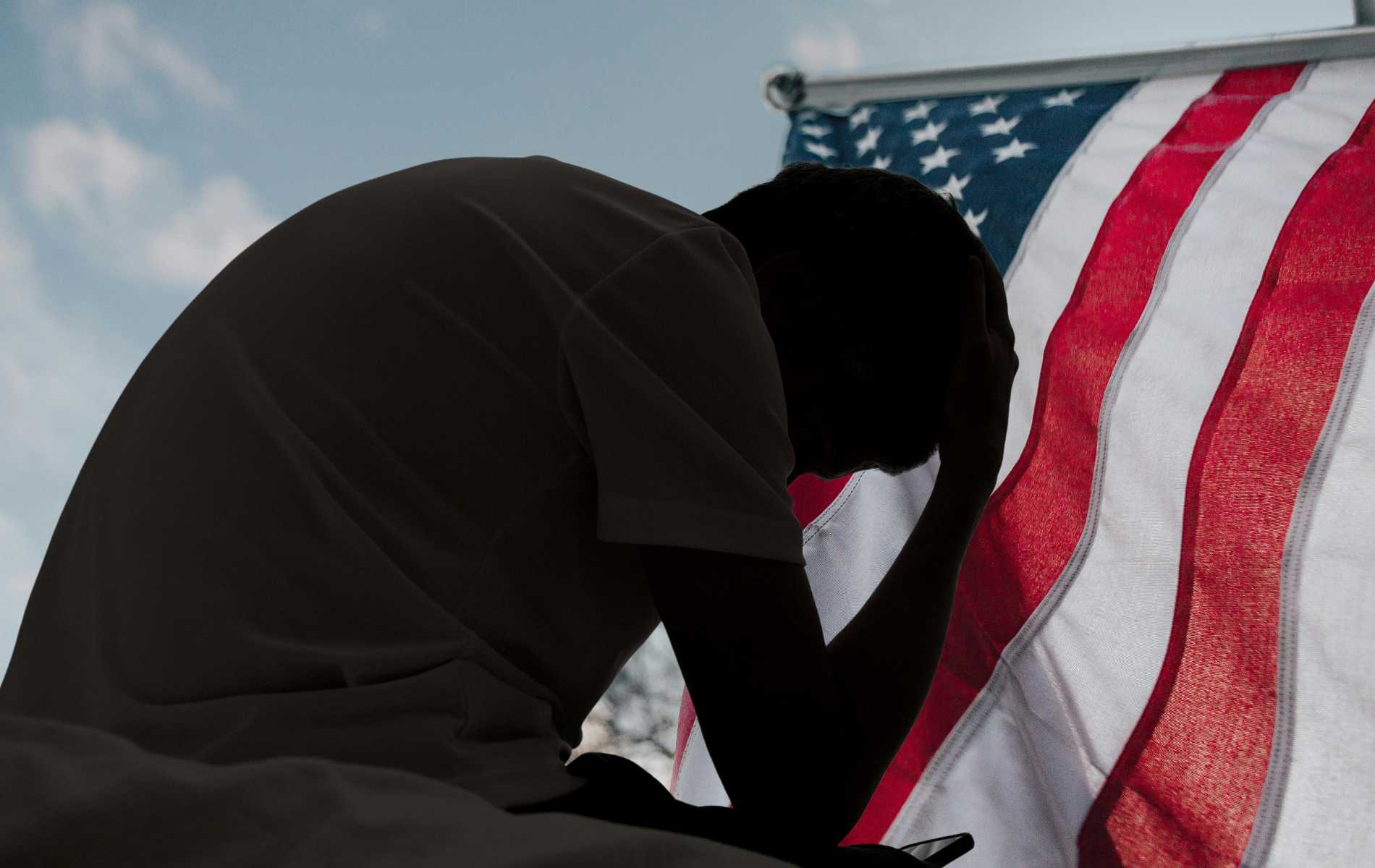 A distressed man in silhouette sits in dark room texting on smartphone. Blinds drawn in window light making silhouette of man with hand of forehead communicating on cell phone. Moody dark image with copy space.