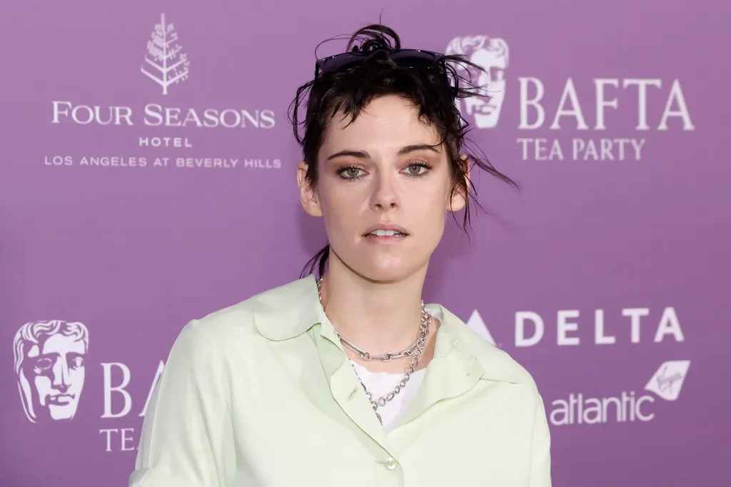 Kristen Stewart con una camisa de color amarillo pálido y dos collares de cadena plateada sobre un fondo violeta de los BAFTA.