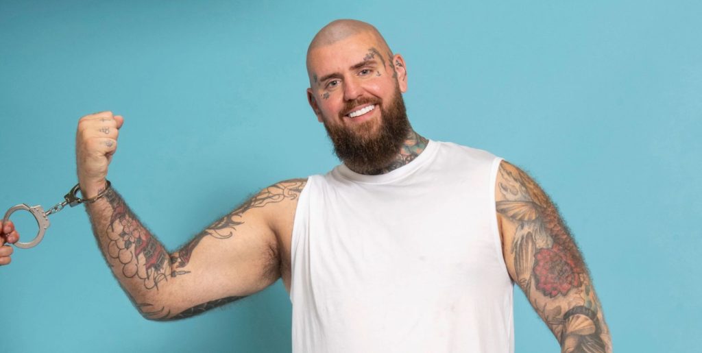 Handcuffed contestant Rob smiling wearing handcuffs while against a blue background.