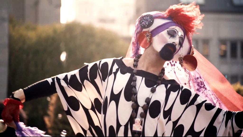 Hunky Jesus todavía: una persona vestida de mujer con un vestido largo blanco y negro y un tocado morado y naranja.