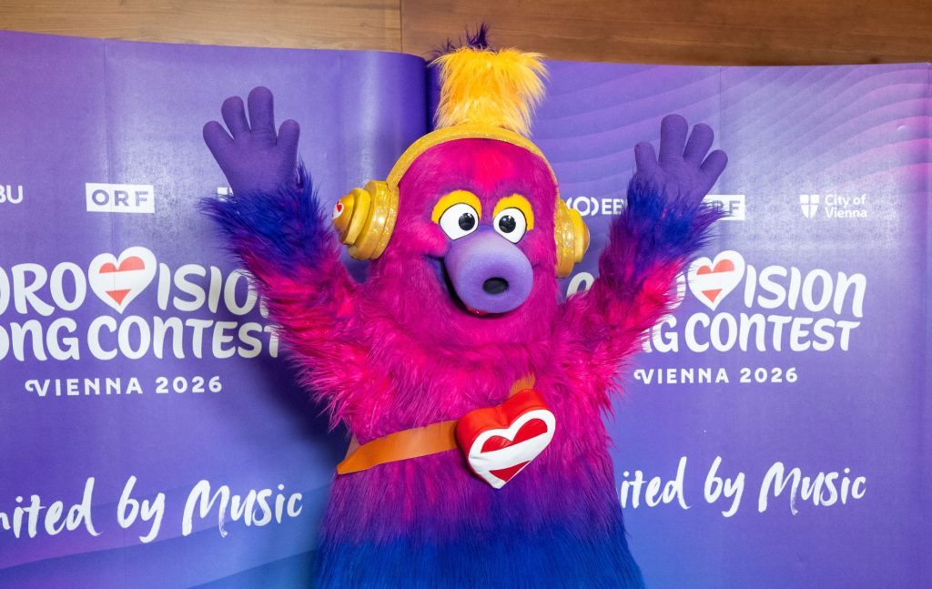 Auri is the mascot of the 2026 Eurovision Song Contest. A fluffy creature which has a pink head that fades to purple and then blue legs. Auri is wearing trainers and has yellow headphones on. Holiday Austria flag heart