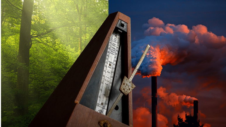 La barra pendular de un metrónomo apunta hacia una escena de chimeneas de fábrica iluminada por el atardecer, mientras que al otro lado del metrónomo se ve una vista iluminada por el sol de árboles verdes.