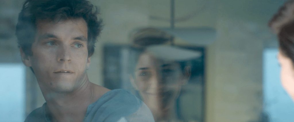 Fionn Whitehead como Philip en Satisfaction. Lleva una blusa en tonos azules y mira por la ventana a una mujer.