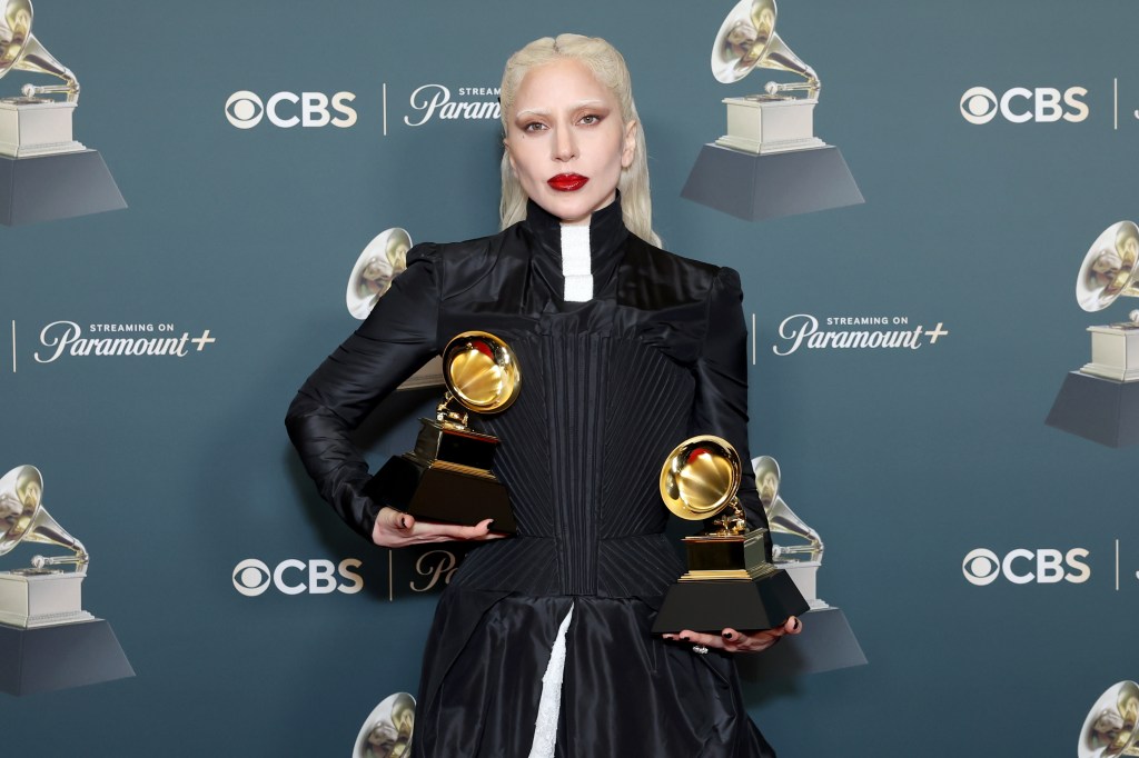 Lady Gaga de pie con dos premios Grammy con un vestido largo negro con detalles blancos debajo.