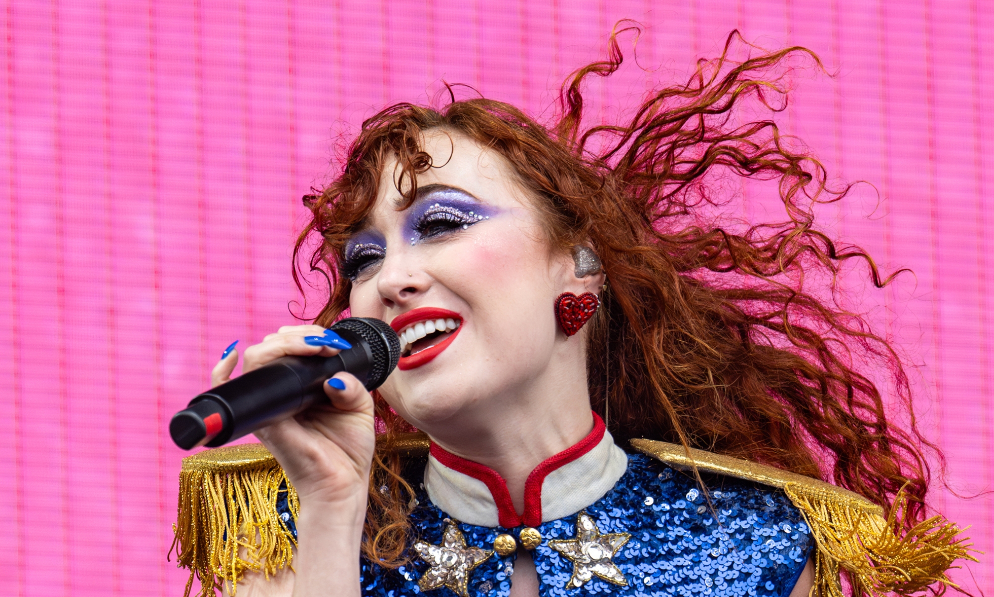 Chappell Roan performing against a bright pink background. She is wearing a blue sequin and gold fringe shirt with big red earrings