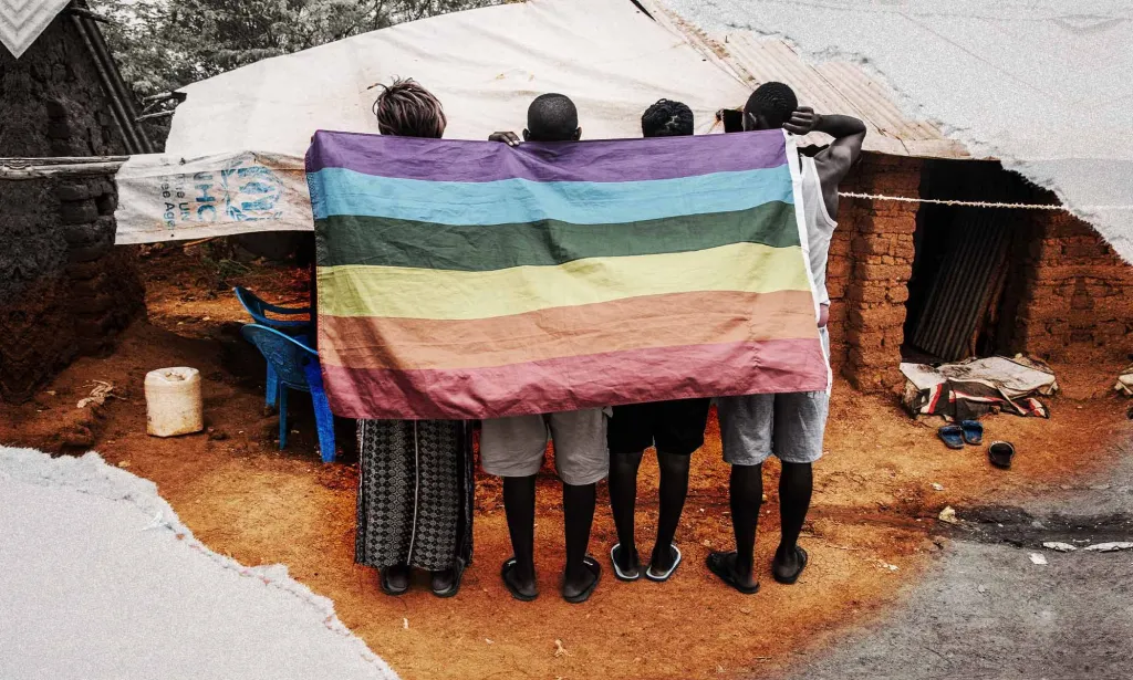 Una imagen muestra a cuatro solicitantes de asilo de pie, de espaldas a la cámara, en el campo de refugiados de Kakuma, Kenia. Llevan una bandera del Orgullo colgada sobre sus espaldas.