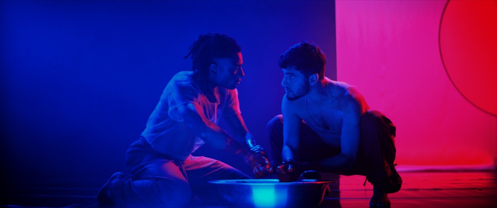 Imagen del escenario nocturno: dos hombres agachados sobre un cuenco, con una luz azul y rosa detrás de ellos.