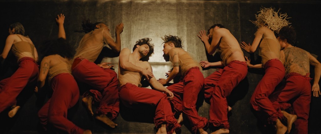 Ocho artistas tumbados sobre un suelo de madera con camisetas de malla color nude y pantalones rojos.