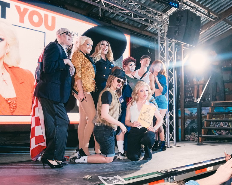 Un grupo de artistas drag aparece en el escenario frente a una gran pantalla de proyección que muestra una imagen promocional del evento.