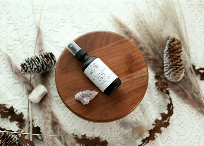 Una botella de Piece of Mynd's Sacred Bliss Magnesium Spray sobre una mesa con piñas y cristales