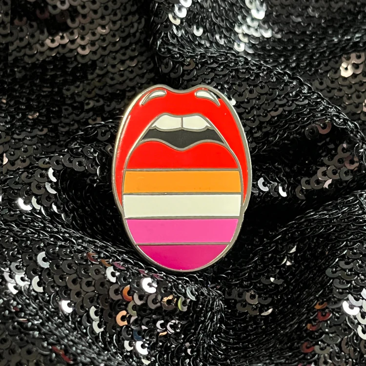 Un pin esmaltado con el diseño de una boca abierta con la lengua afuera y la lengua tiene rayas que se alinean con la bandera del orgullo lésbico.