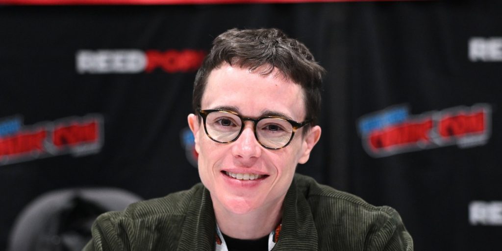 Elliot Page attends New York Comic Con. Eliott wearing a green cordoroy jacket smiling into the camera, wearing round glasses.