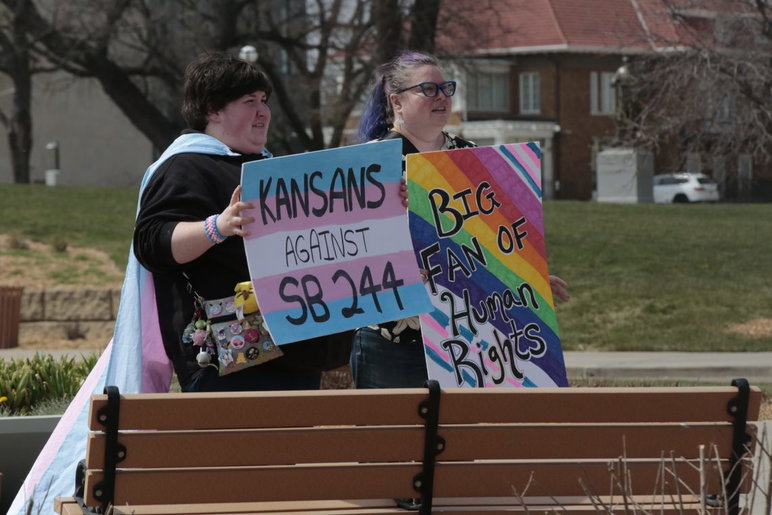 Connor Montgomery, izquierda, y Beth Falkenstein se encuentran afuera del Capitolio de Kansas con carteles para el Día de la Visibilidad Transgénero el 31 de marzo de 2026.