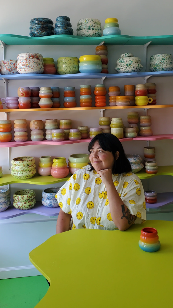 Viviana Matsuda sentada en su estudio frente a un montón de coloridas cerámicas y estantes.
