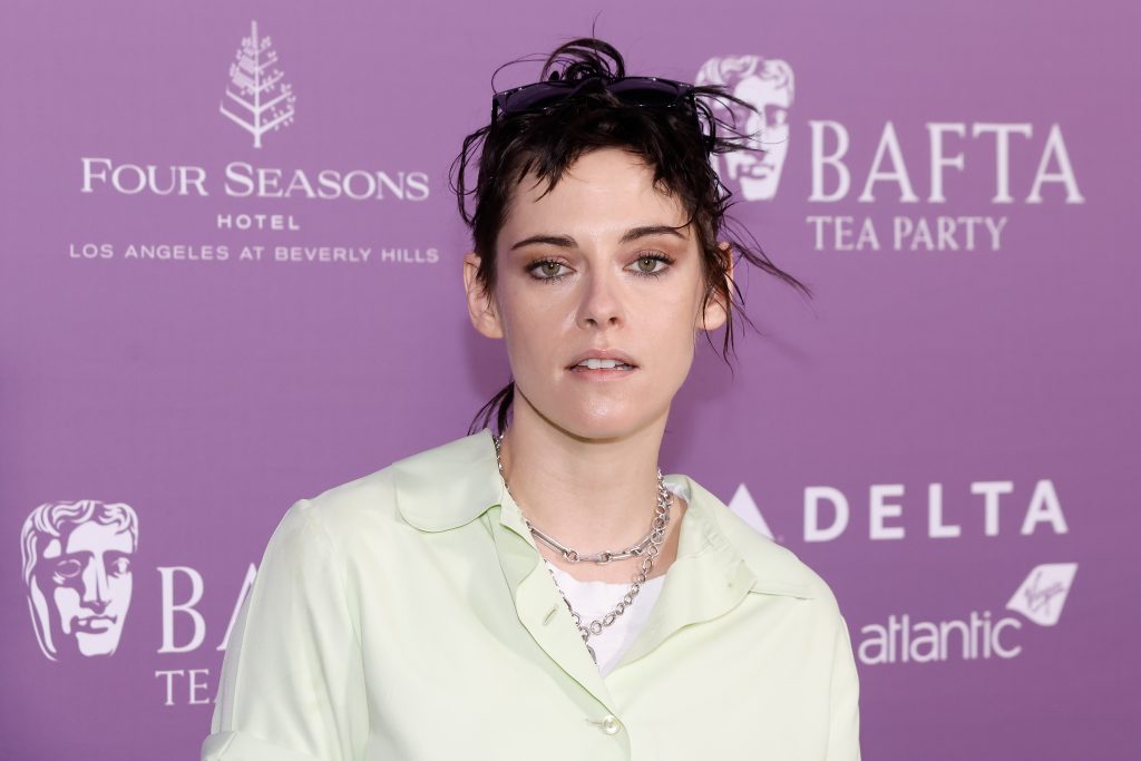 Kristen Stewart in a pale yellow shirt and two silver chain necklaces against a purple BAFTA backdrop