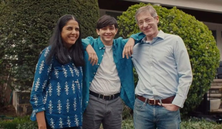 La madre, con cabello largo y negro, tez india y vestido azul con detalles en blanco, está de pie junto a su hijo, un adolescente birracial de cabello negro, una camisa abierta azul, una camiseta blanca y pantalones grises con cinturón negro. El padre de cabello gris y limpio a la derecha del hijo usa una camisa azul pálido con botones, gafas, cinturón y jeans azules con la mano izquierda en el bolsillo. Todos sonríen mientras están parados frente a arbustos verdes durante el día.