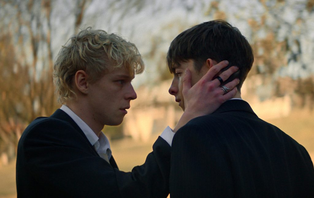 Leviticus still: two teen boys, the blonde cradling the cheek of the brunet, both in suits.
