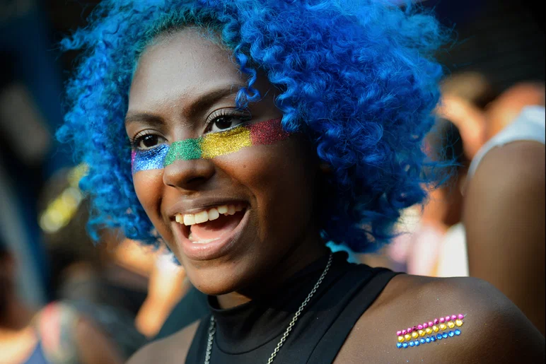 Una joven negra con una peluca azul con volantes sonríe con un brillante maquillaje de arco iris recorriendo sus mejillas y nariz y una bandera del Orgullo Pansexual adornada con pedrería en su hombro izquierdo.