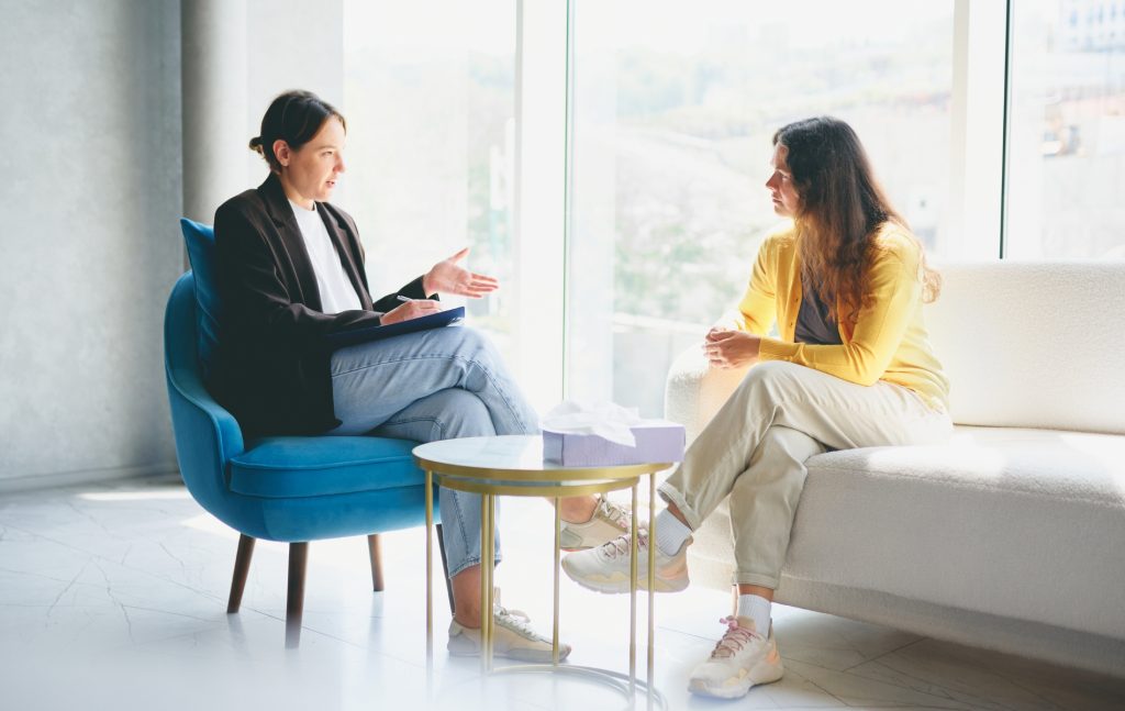 Two women are seated in a bright room with large windows. One woman, wearing a yellow sweater, is listening attentively while the other woman takes notes. The setting is professional, suggesting a counseling or meeting scenario. The room features a blue chair, a white sofa, and a small table with a tissue box. No visible copy space.