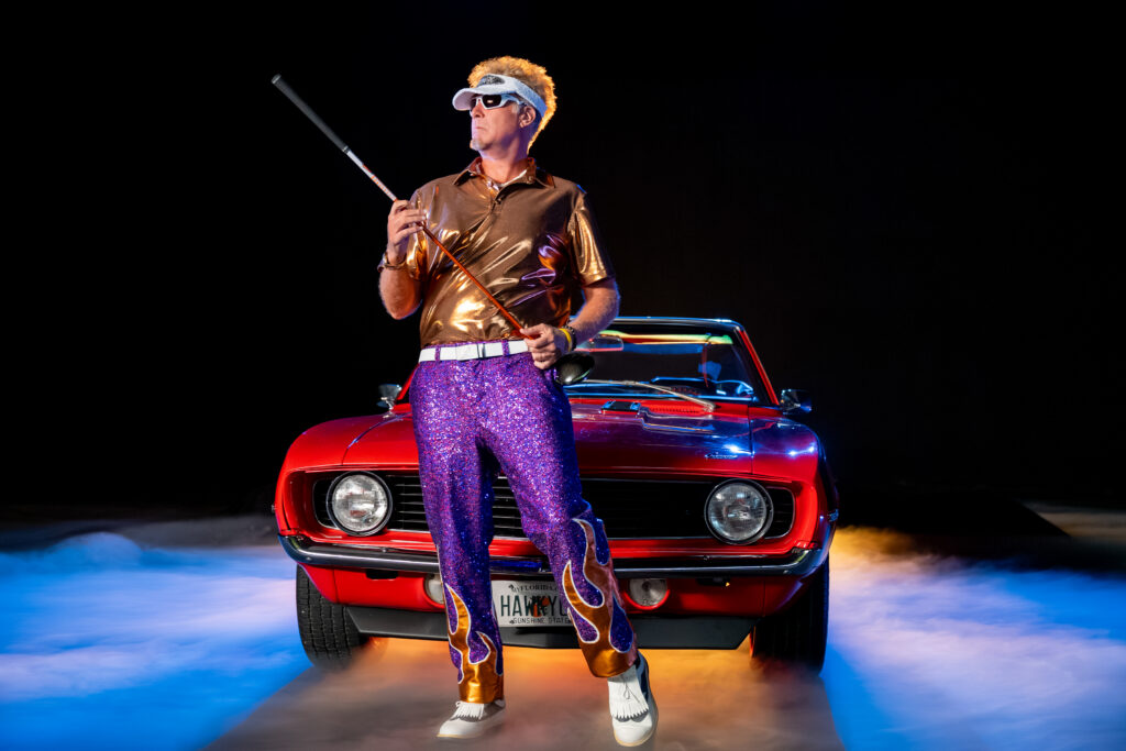 "Hombre con camisa brillante, pantalones de lentejuelas moradas y gafas de sol blancas sosteniendo un bastón, parado frente a un auto convertible vintage rojo con niebla e iluminación colorida, fondo negro, escena dramática y de estilo retro."
